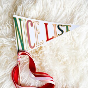 Puede incluir: Un banderín blanco con las palabras "NICE LIST" en letras de colores sobre un fondo blanco esponjoso. El banderín está unido a una varita de cinta roja, blanca y rosa.