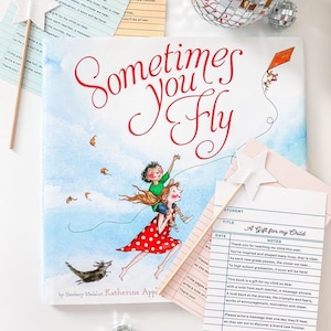 May include: A children's book titled "Sometimes You Fly" with a whimsical illustration of a girl carrying a boy. Accompanying the book are star-shaped decorations, a wand, and a card with text. A disco ball adds a touch of sparkle.