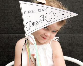 May include: A young girl holds a white pennant with the text "FIRST DAY Pre-K 3" in black lettering. The pennant has a black ribbon with colorful streamers attached.