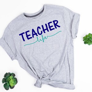 K&ouml;nnte beinhalten: Graues T-Shirt mit dem Text "TEACHER life" in blauer und gr&uuml;ner Schrift.