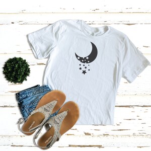 Puede incluir: Camiseta blanca con un gr&aacute;fico de luna creciente negra y estrellas. La camiseta est&aacute; sobre una superficie de madera blanca con un par de sandalias y una peque&ntilde;a planta verde.