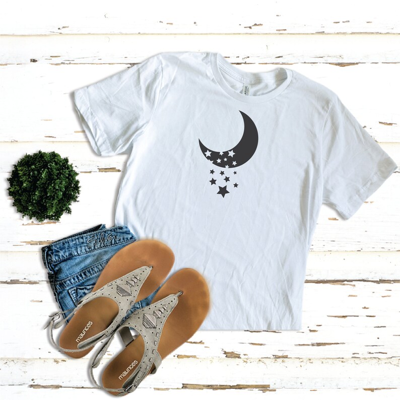 Puede incluir: Camiseta blanca con un gr&aacute;fico de luna creciente negra y estrellas. La camiseta est&aacute; sobre una superficie de madera blanca con un par de sandalias y una peque&ntilde;a planta verde.