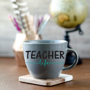 K&ouml;nnte beinhalten: Eine graue Keramiktasse mit dem Text "TEACHER life" in schwarzen Buchstaben und einer blaugr&uuml;nen Unterstreichung. Die Tasse steht auf einer Holzoberfl&auml;che.