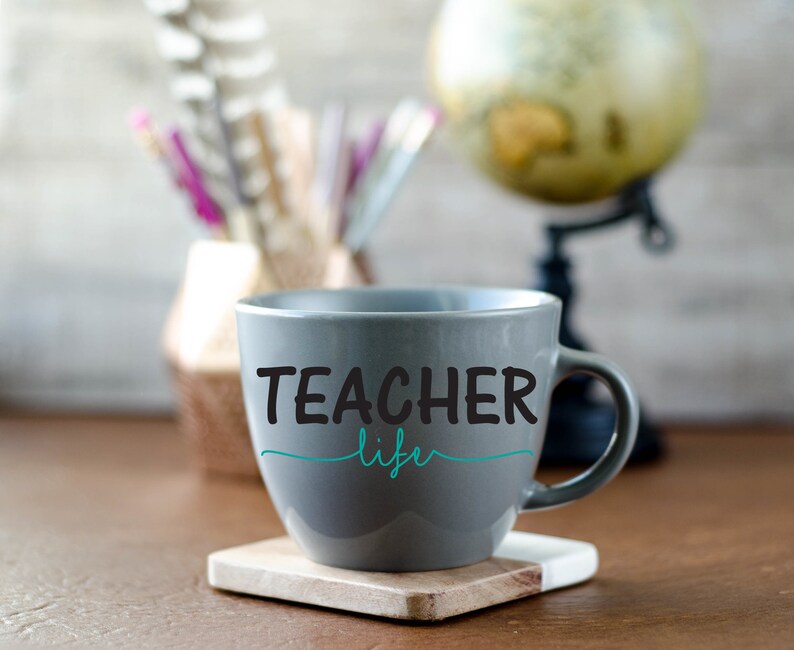 K&ouml;nnte beinhalten: Eine graue Keramiktasse mit dem Text "TEACHER life" in schwarzen Buchstaben und einer blaugr&uuml;nen Unterstreichung. Die Tasse steht auf einer Holzoberfl&auml;che.
