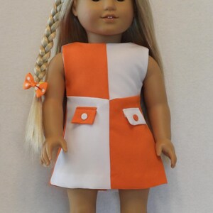 May include: A doll wearing a sleeveless orange and white color-block dress with two small pockets. The doll has long blonde hair in a braid with an orange bow. The doll is wearing white boots.