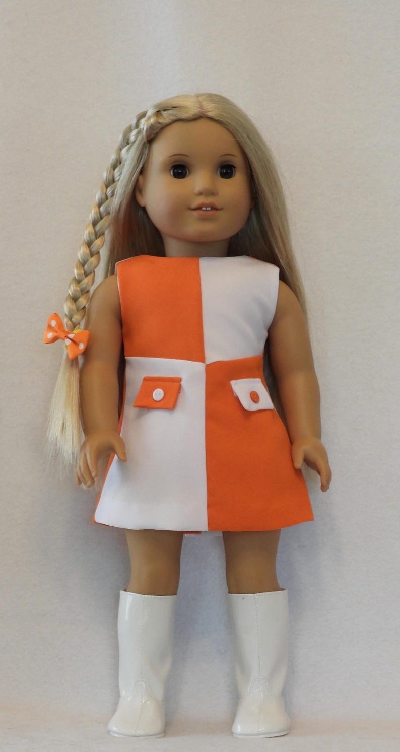 May include: A doll wearing a sleeveless orange and white color-block dress with two small pockets. The doll has long blonde hair in a braid with an orange bow. The doll is wearing white boots.