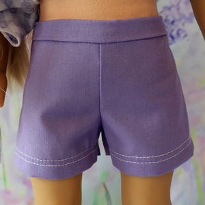 以下が含まれることがあります： 白いステッチが裾に沿って施された、ラベンダー色のショートパンツのクローズアップ。 ショーツは幅広のウエストバンドで、テクスチャード加工された生地で作られています。 ショーツは人形用にデザインされています。