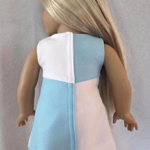 May include: A sleeveless doll dress in a color block design. The dress is divided vertically into three sections: white, light blue, and pink. The doll has long blonde hair.