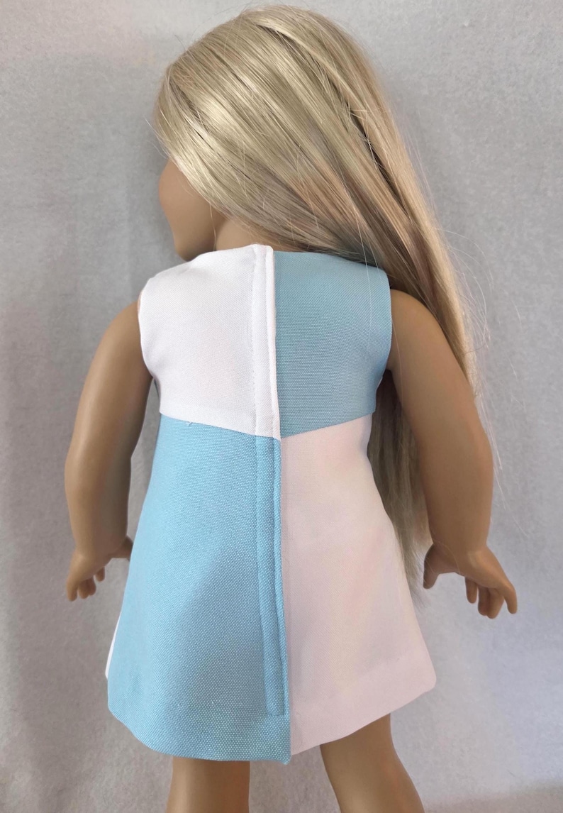 May include: A sleeveless doll dress in a color block design. The dress is divided vertically into three sections: white, light blue, and pink. The doll has long blonde hair.