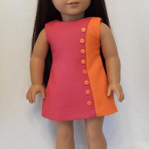 May include: A doll wearing a two-tone dress in shades of pink and orange, fastened with orange buttons. The doll has long, dark hair and is wearing matching orange sandals. The dress is sleeveless and has a simple, A-line cut.
