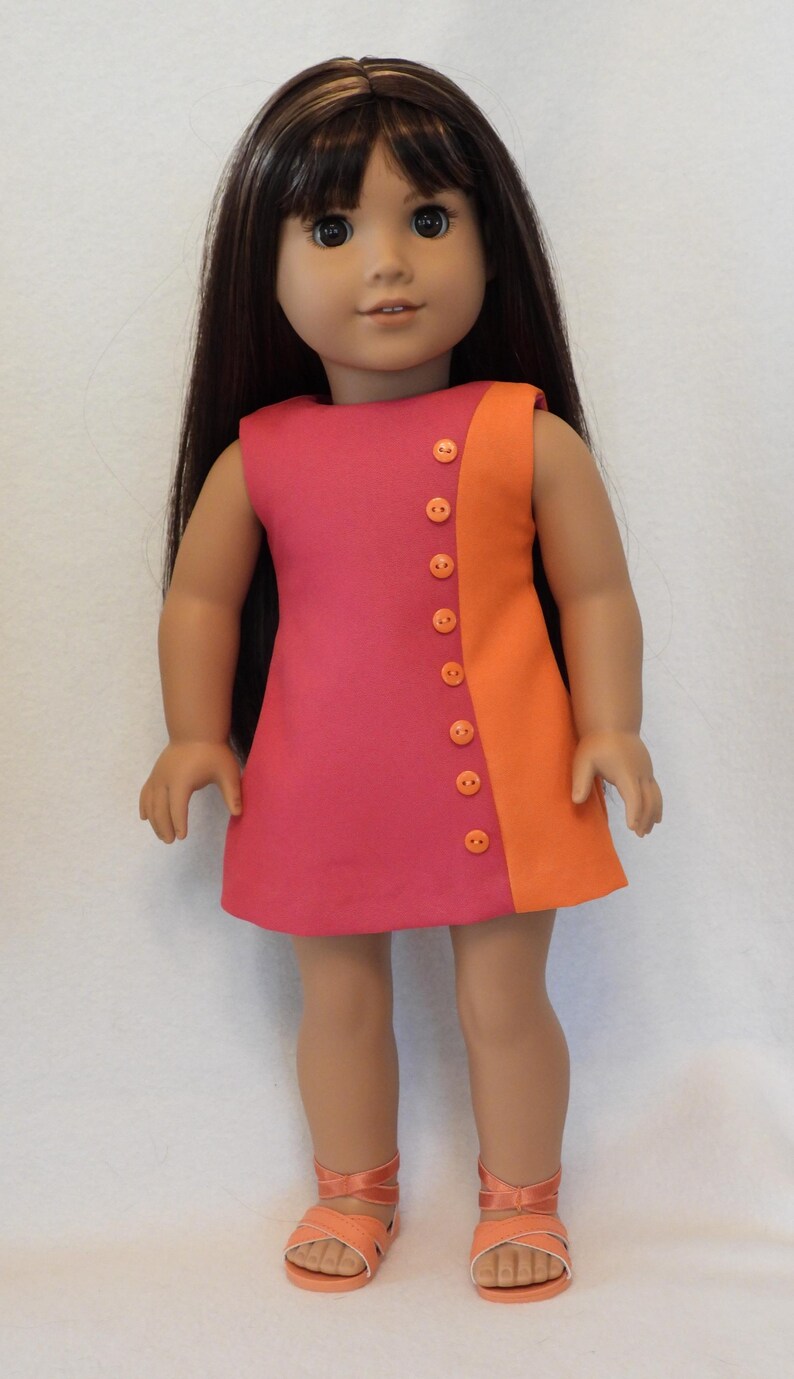 May include: A doll wearing a two-tone dress in shades of pink and orange, fastened with orange buttons. The doll has long, dark hair and is wearing matching orange sandals. The dress is sleeveless and has a simple, A-line cut.