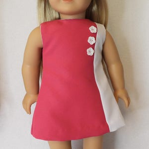 May include: A doll wearing a pink and white dress. The sleeveless dress has a solid pink front and a white panel on the right side. Three white flower-shaped buttons adorn the side. The doll has blonde hair and is wearing pink shoes.