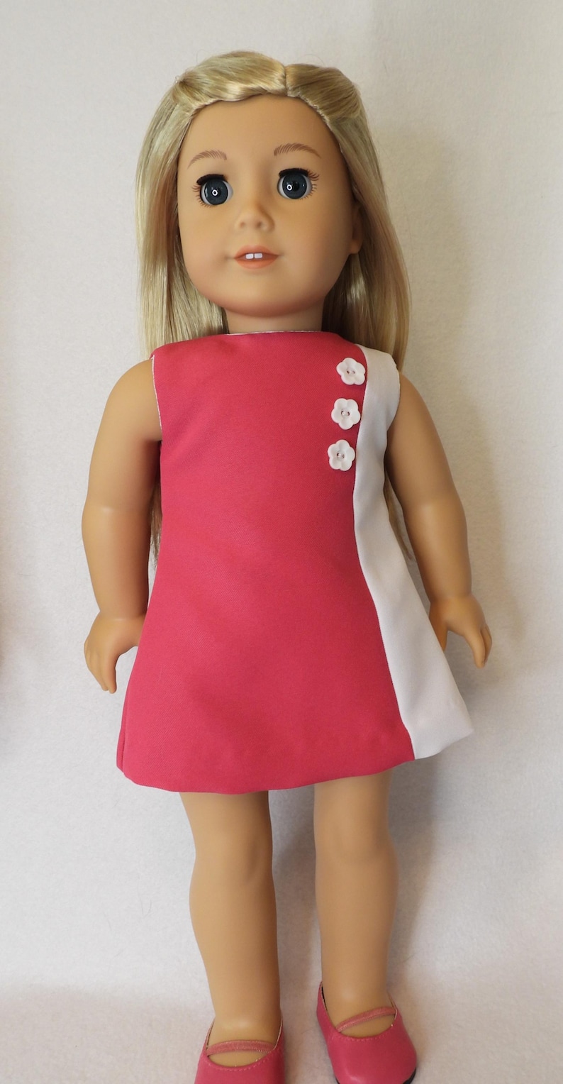May include: A doll wearing a pink and white dress. The sleeveless dress has a solid pink front and a white panel on the right side. Three white flower-shaped buttons adorn the side. The doll has blonde hair and is wearing pink shoes.