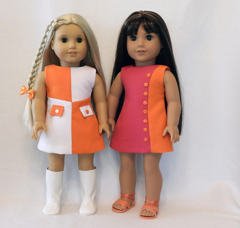 May include: Two dolls wearing colorful dresses. One doll has blonde hair in a braid and is wearing a white and orange dress with white boots. The other doll has dark hair and is wearing a pink and orange dress with orange sandals.