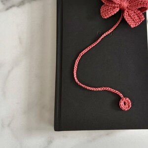 May include: A pink crocheted bow bookmark with a long cord. The bookmark is attached to a black book.