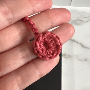May include: A close-up of a pink crocheted circle with a hole in the center. The circle is attached to a pink crocheted cord.