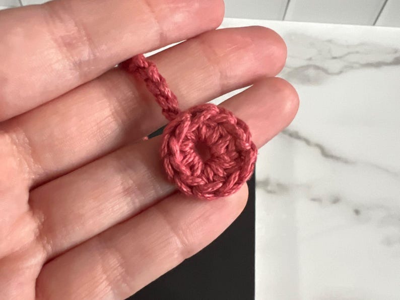 May include: A close-up of a pink crocheted circle with a hole in the center. The circle is attached to a pink crocheted cord.