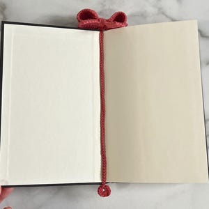 May include: A red crocheted bookmark with a bow at the top and a small circle at the bottom. The bookmark is placed in a closed book.