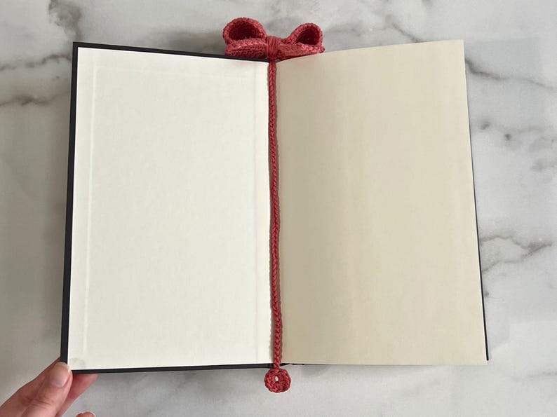 May include: A red crocheted bookmark with a bow at the top and a small circle at the bottom. The bookmark is placed in a closed book.