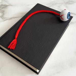 May include: A black book with a white-edged spine is paired with a unique bookmark. The bookmark features a crocheted white eyeball with a blue iris and red veins, connected to a red cord with a tassel.