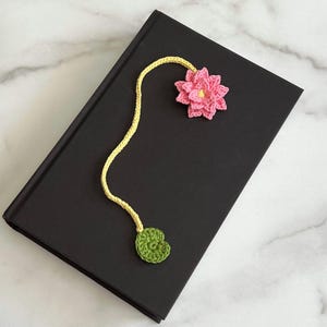 May include: A crocheted bookmark with a pink flower and a green leaf. The bookmark is attached to a black book.