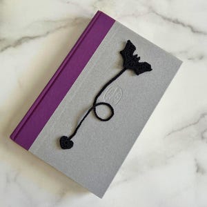 May include: A hardcover book with a purple spine and a light gray cover featuring a black crocheted bookmark. The bookmark consists of a bat-shaped top connected by a thin yarn cord to a small heart-shaped charm at the bottom end.