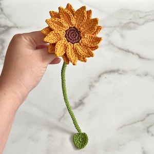 Pode incluir: Um girassol de crochê com um centro marrom e pétalas amarelas. O girassol está preso a um caule verde com uma única folha verde.