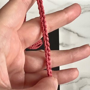 May include: A close-up of a pink braided cord held in a hand. The cord is made of a soft, pliable material and has a smooth, even texture.