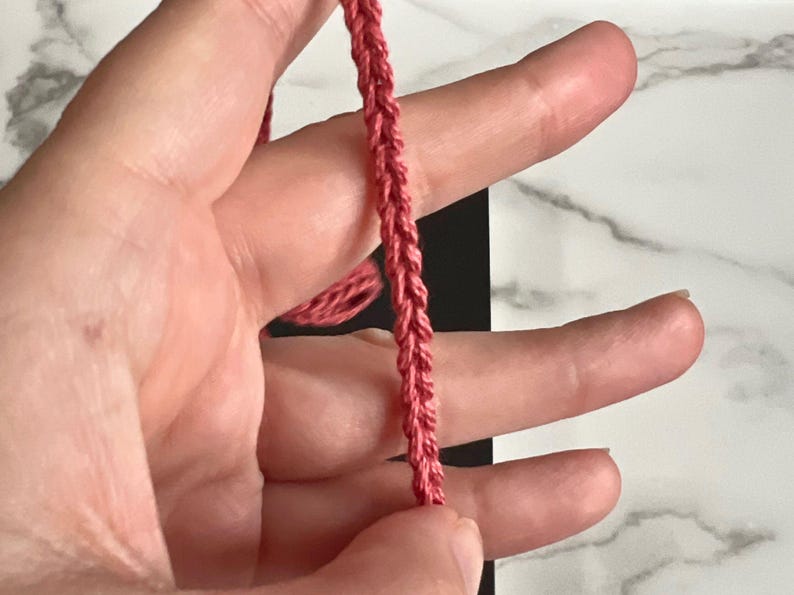 May include: A close-up of a pink braided cord held in a hand. The cord is made of a soft, pliable material and has a smooth, even texture.
