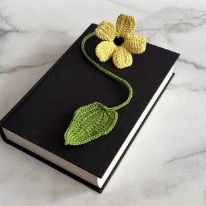 May include: A handmade crochet bookmark featuring a yellow flower and green leaf. The bookmark is placed on a closed black book. The flower and leaf are connected by a green stem. The background is a marble surface.