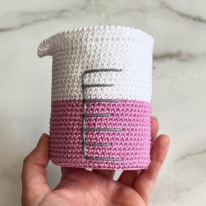 Puede incluir: Un vaso de precipitados de ganchillo hecho a mano en blanco y rosa, con marcas de medición grises. La mitad superior es blanca y la inferior es rosa. El vaso tiene un pequeño pico y está diseñado para parecerse a un vaso de laboratorio.
