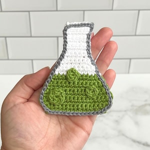 Op de afbeelding: Een handgemaakte gehaakte erlenmeyer in wit en groen, met een grijze rand. De erlenmeyer heeft een witte bovenkant en een groene basis met drie decoratieve cirkelvormige elementen. Het item wordt in een hand gehouden.