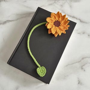 May include: A crocheted sunflower bookmark with a green stem and heart-shaped leaf. The bookmark is on a black book.