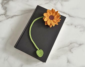 May include: A crocheted sunflower bookmark with a green stem and heart-shaped leaf. The bookmark is on a black book.