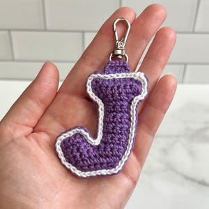 May include: A purple crochet letter J keychain featuring a white stitched border and a silver-toned metal lobster clasp. The letter is crafted with a textured yarn finish and is shaped in a bold, block-style font with rounded edges.