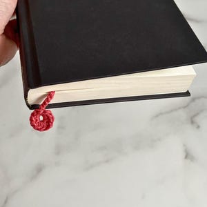 May include: A black hardcover book with a red crocheted bookmark. The bookmark is tied in a circle.