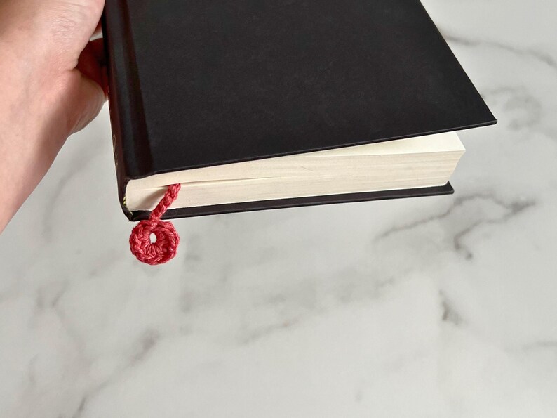 May include: A black hardcover book with a red crocheted bookmark. The bookmark is tied in a circle.