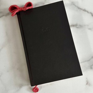 May include: A black hardcover book with a red crocheted bow bookmark. The book has a small embossed logo on the front cover.
