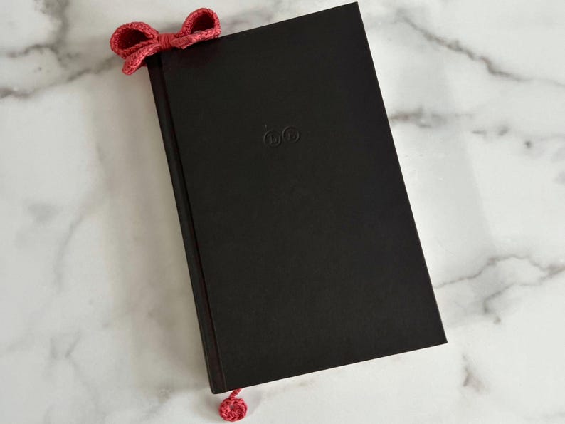 May include: A black hardcover book with a red crocheted bow bookmark. The book has a small embossed logo on the front cover.