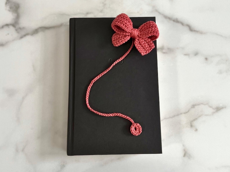 May include: A pink crocheted bow bookmark with a long cord. The bookmark is attached to a black book.