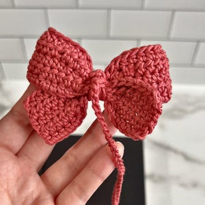 May include: A crocheted bow made of pink yarn. The bow is tied with a long tail of yarn.