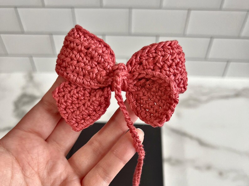 May include: A crocheted bow made of pink yarn. The bow is tied with a long tail of yarn.