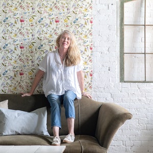 May include: A woman sits on a green velvet sofa in front of a white brick wall. She is wearing a white shirt, blue jeans, and white sneakers. The wall behind her is covered in a floral wallpaper with a white background and colorful flowers. The wallpaper is labeled "Pasted Paper" and "PrePasted Paper Sheets for Covering Your Walls & More". The sofa is in front of a white table with a white top and wooden legs. The V&A logo is visible in the bottom right corner of the image.