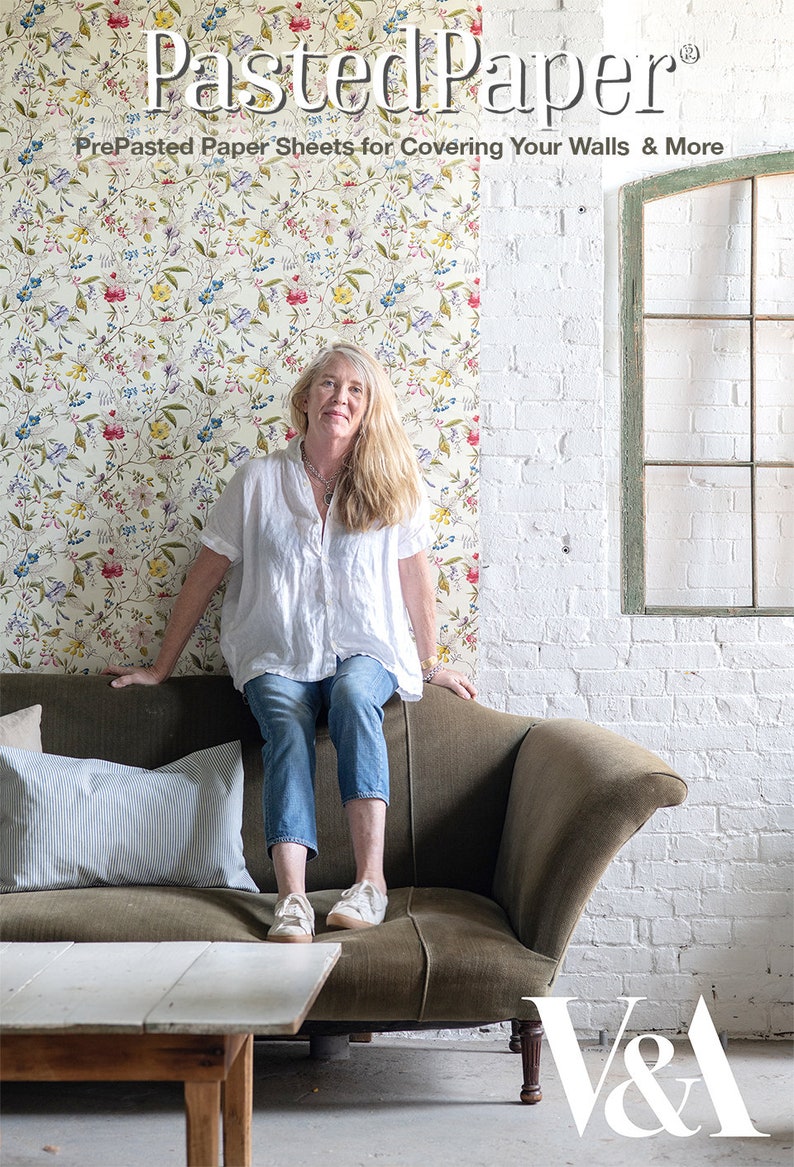 May include: A woman sits on a green velvet sofa in front of a white brick wall. She is wearing a white shirt, blue jeans, and white sneakers. The wall behind her is covered in a floral wallpaper with a white background and colorful flowers. The wallpaper is labeled "Pasted Paper" and "PrePasted Paper Sheets for Covering Your Walls & More". The sofa is in front of a white table with a white top and wooden legs. The V&A logo is visible in the bottom right corner of the image.