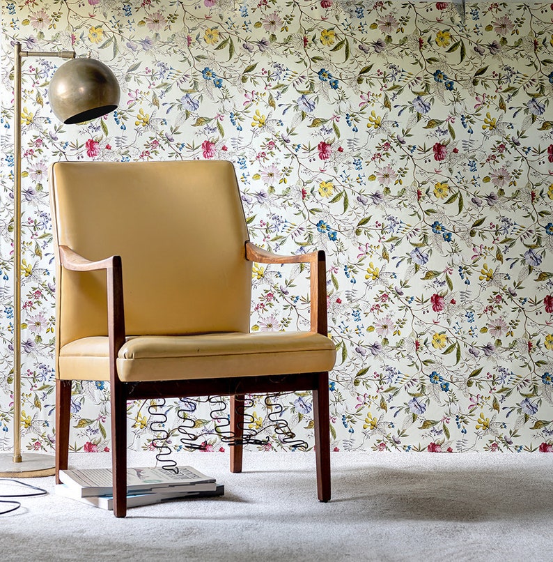 May include: A tan armchair with wooden arms and legs sits in front of a wall covered in a floral wallpaper with a white background. The wallpaper features a repeating pattern of small pink, blue, and yellow flowers.