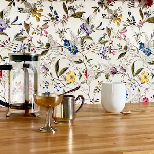 May include: A glass French press coffee maker with a black handle, a silver creamer, a gold-colored metal cup, and a white ceramic mug on a wooden countertop. The background is a white floral wallpaper with a variety of colorful flowers.