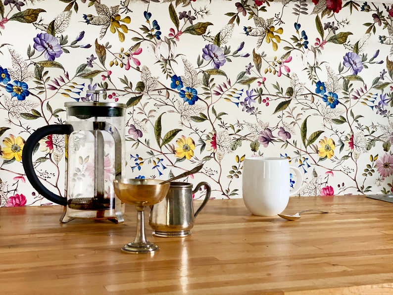May include: A glass French press coffee maker with a black handle, a silver creamer, a gold-colored metal cup, and a white ceramic mug on a wooden countertop. The background is a white floral wallpaper with a variety of colorful flowers.