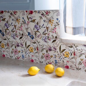 May include: A close-up of a kitchen backsplash with a floral pattern in shades of pink, purple, yellow, and green. Three yellow lemons are sitting on a white countertop in front of the backsplash.