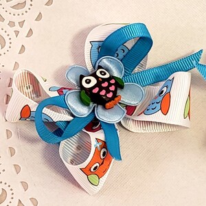 May include: A handmade hair bow featuring a central owl embellishment. The bow is made of white ribbon printed with cartoon owls in orange, blue, and green. A blue ribbon is looped around the bow, and a small owl is in the center.