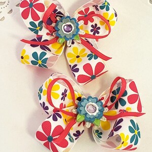 May include: Two colorful hair bows with a white background and a floral pattern. Each bow has a red ribbon and a blue and green flower with a sparkly center.
